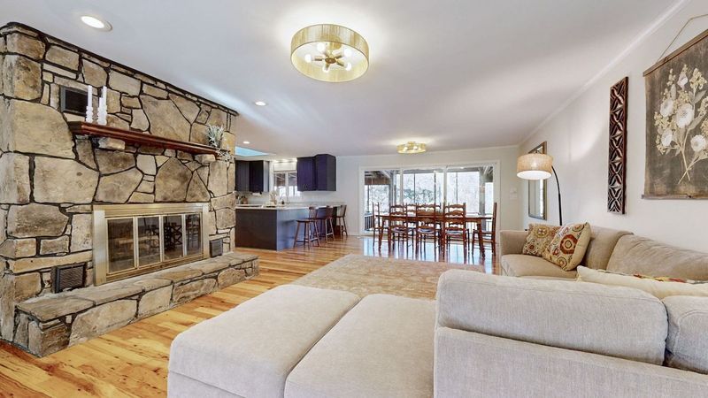 Main-level living space featuring a stone fireplace, cozy seating, and hardwood floors. Opens to a fully equipped kitchen and dining area with forest views—an inviting space for gathering, relaxing, and enjoying comfortable cabin-style living.